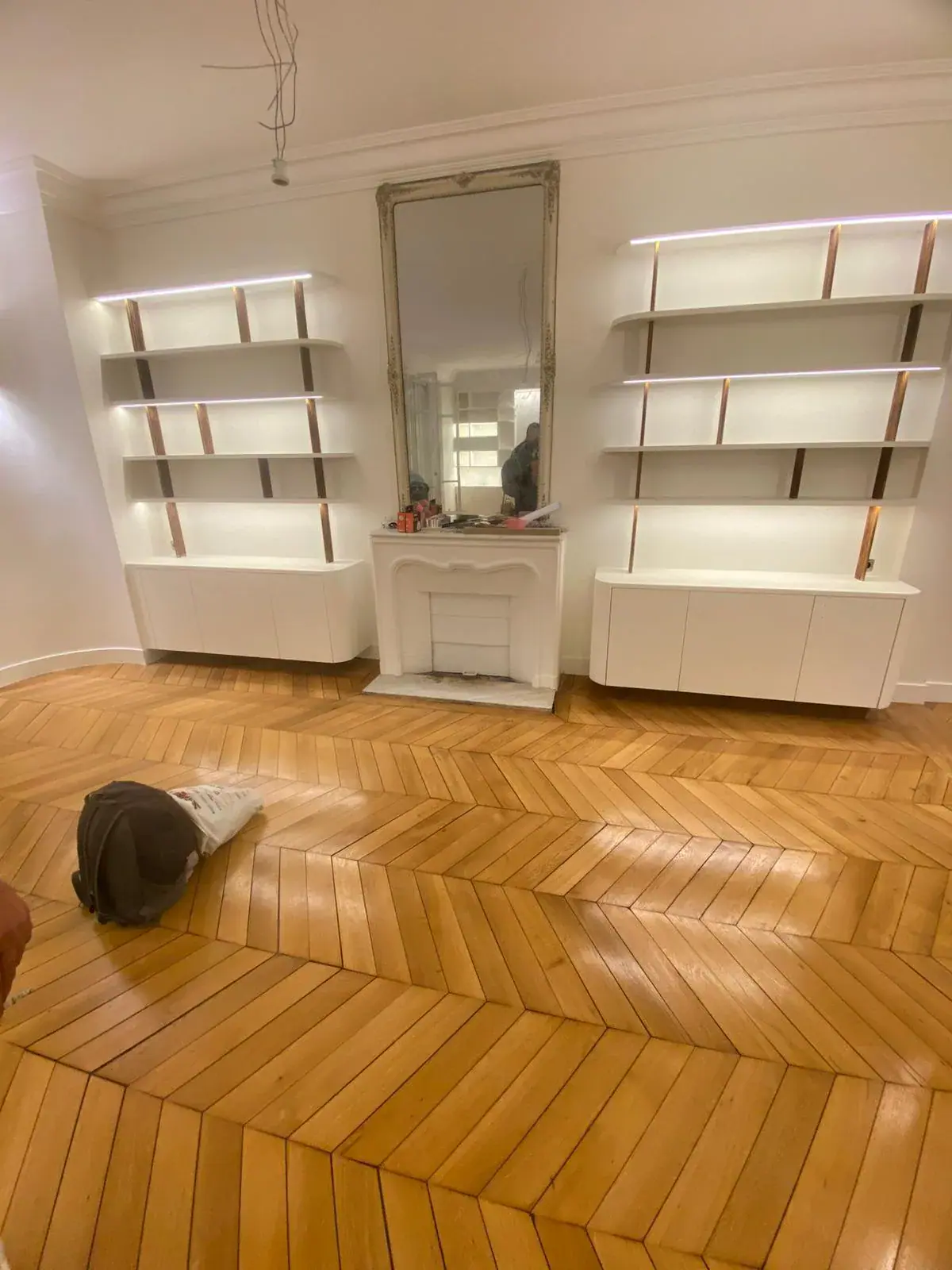 Bibliothèques blanches LED et cheminée avec miroir doré sur parquet chevron — menuiserie Les Artistes